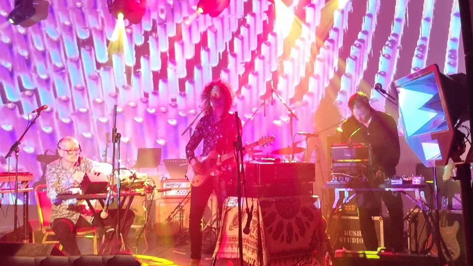 A three-piece band performing on a lit stage, with synthesizers, a laptop and an electric guitar set up on tables and stands.