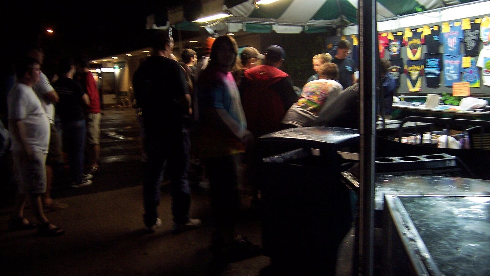 A night-time merch stand at an outdoor concert, with T-shirts hung on the back wall and fans queueing at the counter.